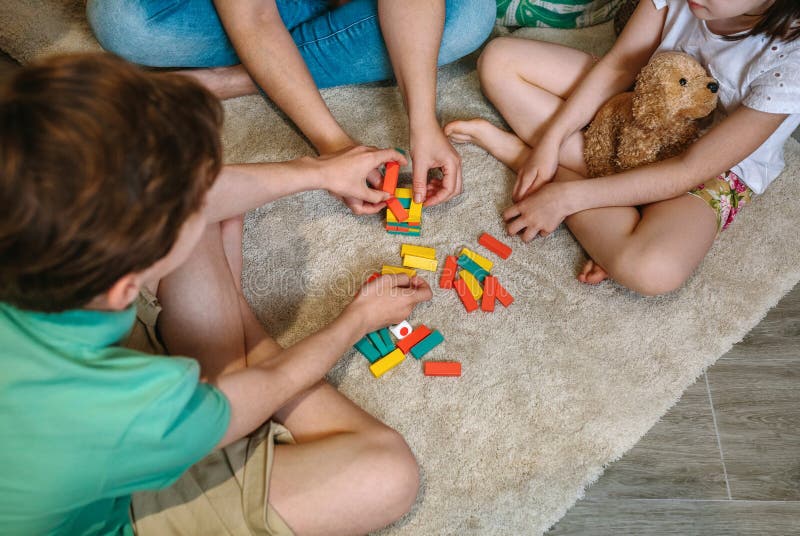 Unrecognizable mother and sons having fun playing wooden stacking piece game over carpet at home. Single parent family enjoying together in leisure time. Happy little girl holding stuffed teddy dog. Unrecognizable mother and sons having fun playing wooden stacking piece game over carpet at home. Single parent family enjoying together in leisure time. Happy little girl holding stuffed teddy dog.