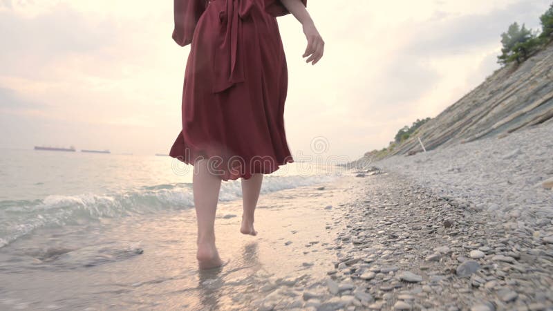 Ноги молодой девушки с красным платье из-за спины ходят по скалистому пляжу на морском побережье на закате Платье расцветает