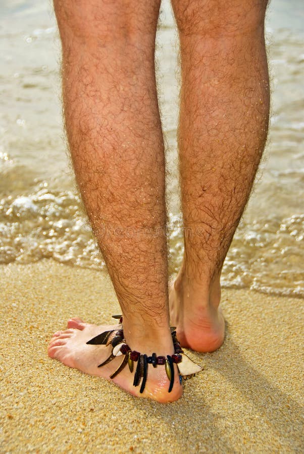 Hairy legs of man with ankle bracelet stood on sand with sea in background. Hairy legs of man with ankle bracelet stood on sand with sea in background.