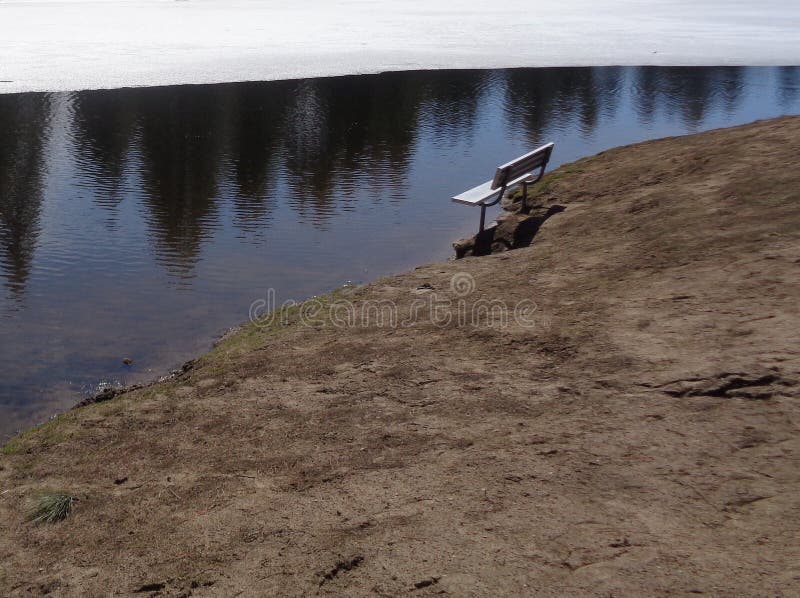 A vacant bench stands on the edge of a winter frozen lake. The aluminum bench is standing in the mud next to a partially frozen lake. The cold lake water reflects the opposite shoreline with pine trees and blue sky. The ground beneath the bench is wet mud, which extends down to the muddy lake bottom in front of the bench. The colors are muted tones of white, silver, blue and brown. The mood is somber and quiet. This image could be used to represent feelings of loneliness, depression, melancholy and isolation.  It could also be used to express SAD Seasonal Affective Disorder, grief, sadness, loneliness, depression, or anxiety.   This image could be used as a background or screensaver.  There is room for text at the top. A vacant bench stands on the edge of a winter frozen lake. The aluminum bench is standing in the mud next to a partially frozen lake. The cold lake water reflects the opposite shoreline with pine trees and blue sky. The ground beneath the bench is wet mud, which extends down to the muddy lake bottom in front of the bench. The colors are muted tones of white, silver, blue and brown. The mood is somber and quiet. This image could be used to represent feelings of loneliness, depression, melancholy and isolation.  It could also be used to express SAD Seasonal Affective Disorder, grief, sadness, loneliness, depression, or anxiety.   This image could be used as a background or screensaver.  There is room for text at the top.