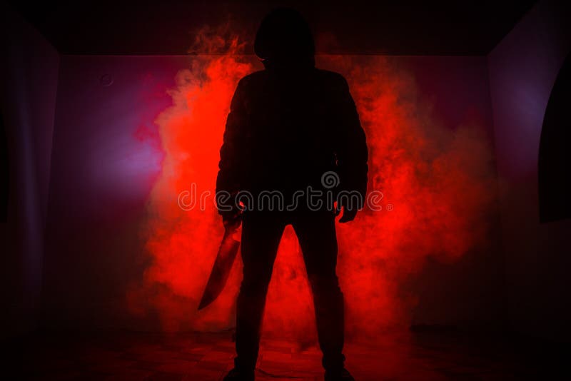 A dangerous hooded man standing in the dark and holding a knife. Face can not be seen. Committing a crime concept. A dangerous hooded man standing in the dark and holding a knife. Face can not be seen. Committing a crime concept