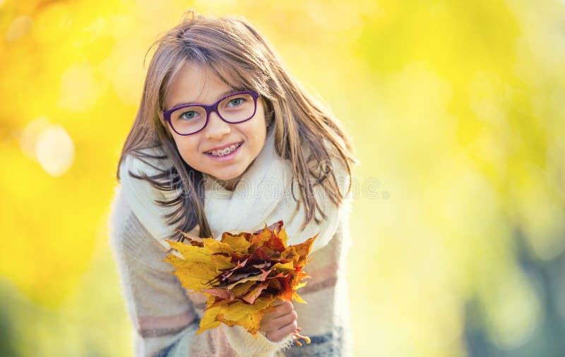 Autumn. Portrait of a smiling young girl who is holding in her hand a bouquet of autumn maple leaves.Pre-teen young girl with glasses and teeth braces. Autumn. Portrait of a smiling young girl who is holding in her hand a bouquet of autumn maple leaves.Pre-teen young girl with glasses and teeth braces.