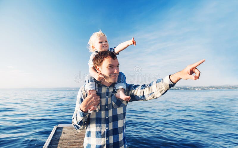 Father and son on sea and sky backgrounds. Little boy is sitting on father`s shoulders and points forward. Parent and child together at summer. Family, Lifestyle, Holidays and Travel concept. Father and son on sea and sky backgrounds. Little boy is sitting on father`s shoulders and points forward. Parent and child together at summer. Family, Lifestyle, Holidays and Travel concept.