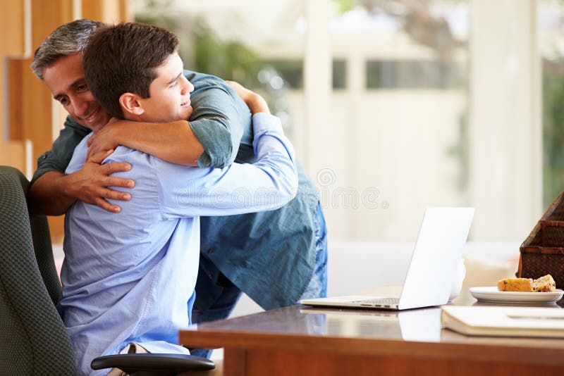 Father And Teenage Son Having A Hug At Home With Laptop On Table. Father And Teenage Son Having A Hug At Home With Laptop On Table