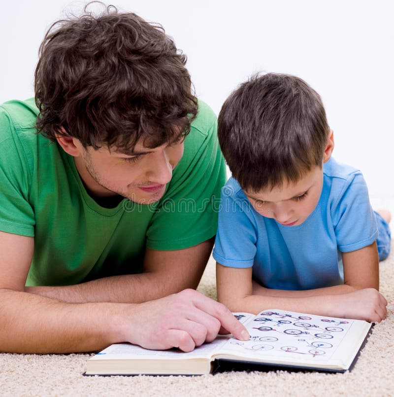 Father and son indoors reading book lying on floor. Father and son indoors reading book lying on floor
