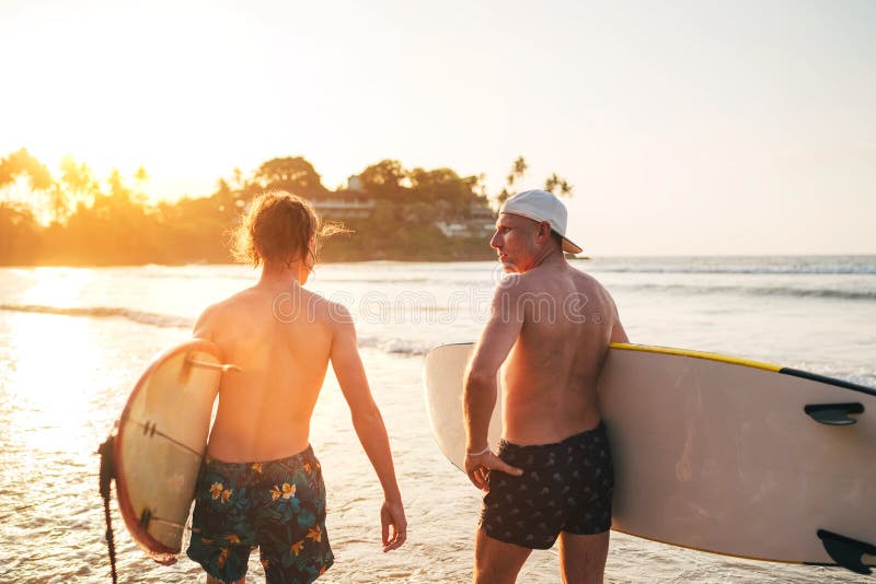 Father with teenager son walking with surfboards by sandy ocean beach with palm trees on background lightened with sunset sun They have conversation and enjoy summertime Family active vacation concept. Father with teenager son walking with surfboards by sandy ocean beach with palm trees on background lightened with sunset sun They have conversation and enjoy summertime Family active vacation concept