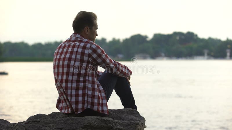Отжатый взрослый мужчина сидя на береге реки и думая о разводе, одиночестве