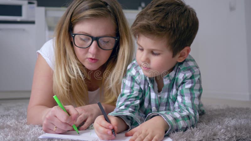 отношение Мат-ребенка, женское с мальчиком с покрашенными карандашами рисует изображения на листе лежа на поле в комнате
