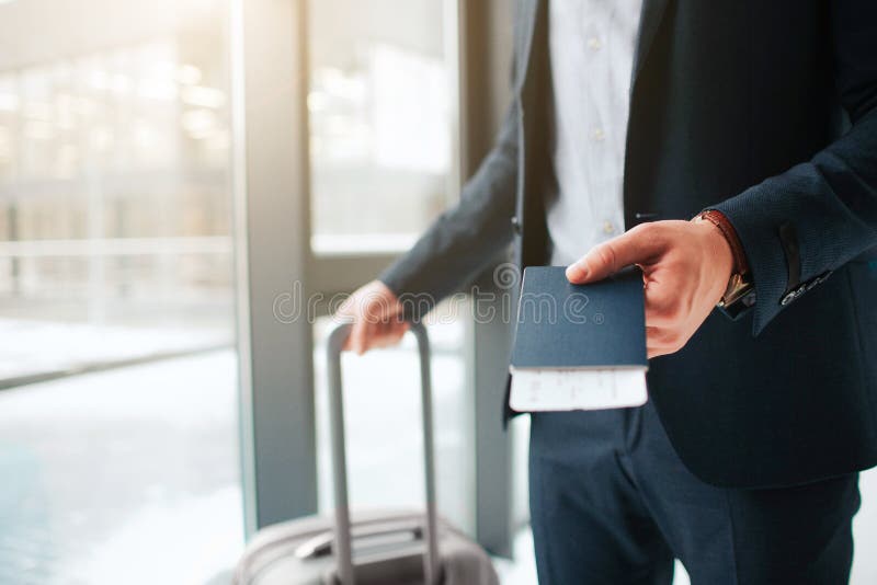 Отрежьте взгляд человека в стойке костюма на окне в аэропорте Он держит паспорт с билетом в нем и чемодан с другой рукой стоковые изображения rf Отрежьте взгляд человека в стойке костюма на окне в аэропорте Он держит паспорт с билетом в нем и чемодан с другой рукой стоковые изображения rf