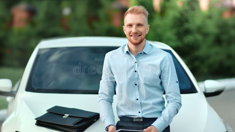 Очаровывая счастливый положительный молодой бизнесмен представляя на открытом воздухе смотря камеру сидя на bonnet автомобиля
