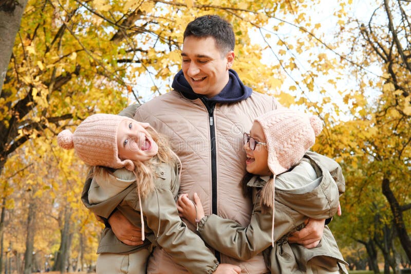 Dad and twin sisters have fun in the autumn park. Dad took the girls under the armpits. Horizontal photo. Dad and twin sisters have fun in the autumn park. Dad took the girls under the armpits. Horizontal photo