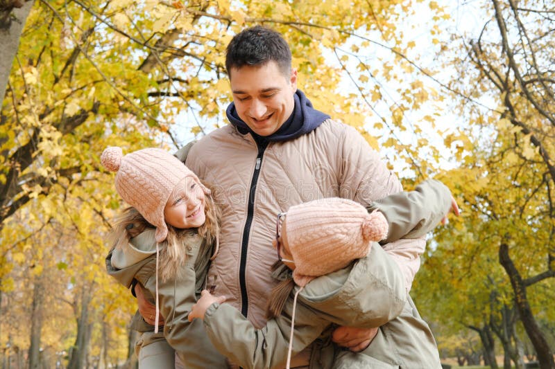 Dad and twin sisters have fun in the autumn park. Dad took the girls under the armpits. Horizontal photo. Dad and twin sisters have fun in the autumn park. Dad took the girls under the armpits. Horizontal photo