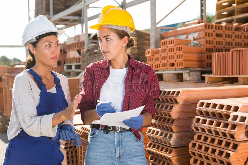 Young woman - manager discussing order list with asian female worker at warehouse of building materials. Young woman - manager discussing order list with asian female worker at warehouse of building materials