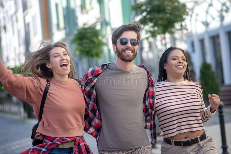 Happy moment. Guy in plaid shirt with sun glasses and two pretty long-haired girls walking enthusiastic laughing with open mouths. Happy moment. Guy in plaid shirt with sun glasses and two pretty long-haired girls walking enthusiastic laughing with open mouths