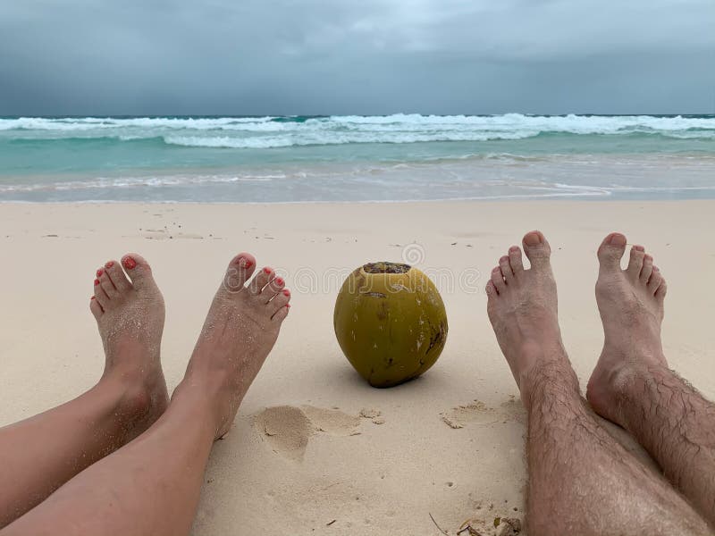 Two pairs of female and male legs with a coconut between them sitting on a white sand Caribbean beach on the ocean background on a cloudy day. Honeymoon image concept. Two pairs of female and male legs with a coconut between them sitting on a white sand Caribbean beach on the ocean background on a cloudy day. Honeymoon image concept