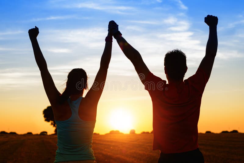 Successful couple of young athletes raising arms to golden summer sunset sky after training. Fitness men and women with arms up celebrating sport goals after exercising in countryside field. Successful couple of young athletes raising arms to golden summer sunset sky after training. Fitness men and women with arms up celebrating sport goals after exercising in countryside field.