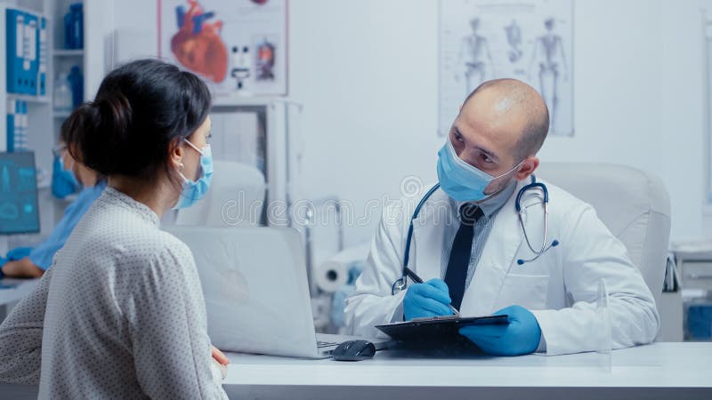 Doctor questioning patient during covid-19 pandemic, writing answears in clipboard. Medical consultation in protective equipment concept shot of sars-cov-2 global health pandemic crisis. Doctor questioning patient during covid-19 pandemic, writing answears in clipboard. Medical consultation in protective equipment concept shot of sars-cov-2 global health pandemic crisis