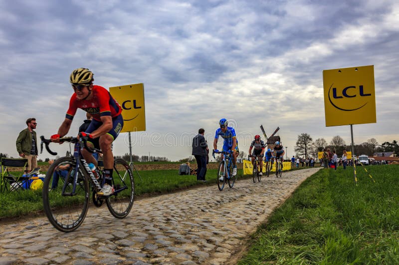 Templeuve, France - April 08, 2018: The peloton riding on the cobblestone road in Templeuve in front of the traditional Vertain Windmill during Paris-Roubaix 2018. Templeuve, France - April 08, 2018: The peloton riding on the cobblestone road in Templeuve in front of the traditional Vertain Windmill during Paris-Roubaix 2018