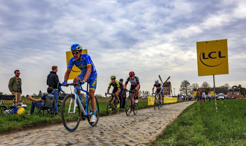 Templeuve, France - April 08, 2018: The peloton riding on the cobblestone road in Templeuve in front of the traditional Vertain Windmill during Paris-Roubaix 2018. Templeuve, France - April 08, 2018: The peloton riding on the cobblestone road in Templeuve in front of the traditional Vertain Windmill during Paris-Roubaix 2018