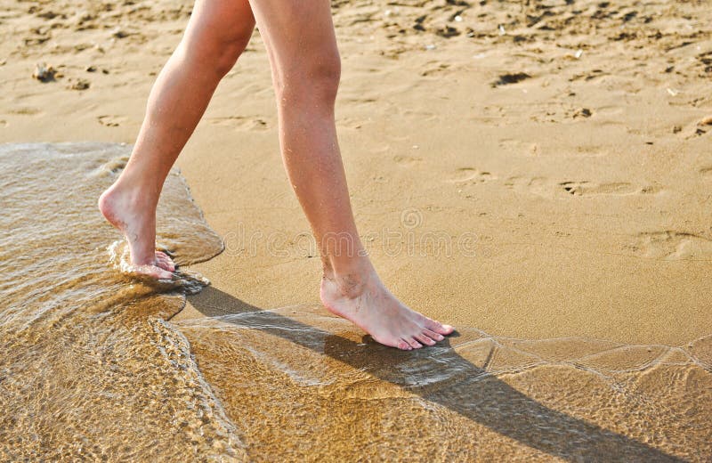 Beach travel - young girl walking on sand beach leaving footprints in the sand. Closeup detail of female feet and golden sand. Teenage female,walking down the beach. Nice legs walking near water. Beach travel - young girl walking on sand beach leaving footprints in the sand. Closeup detail of female feet and golden sand. Teenage female,walking down the beach. Nice legs walking near water