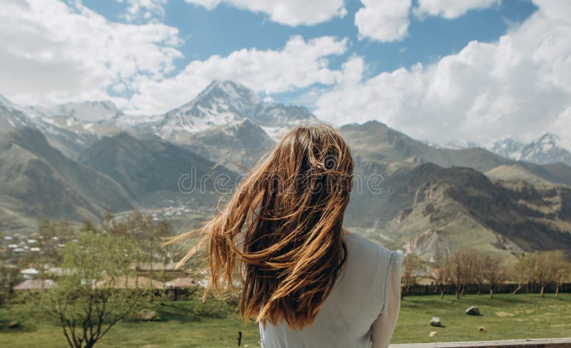 Girl in a dress behind in the summer looks at the mountains with snow peaks. Girl in a dress behind in the summer looks at the mountains with snow peaks