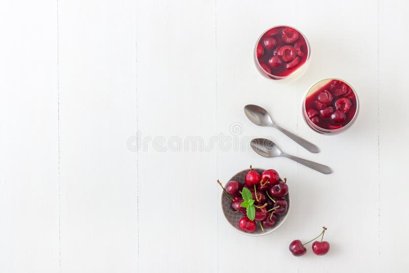 Panna cotta Italian traditional dessert with cherries poached in rum and honey syrup. The bowl of the fresh cherries and 2 glass of panna cotta with spoons. White wooden table background. Top down view. Copy space. Panna cotta Italian traditional dessert with cherries poached in rum and honey syrup. The bowl of the fresh cherries and 2 glass of panna cotta with spoons. White wooden table background. Top down view. Copy space