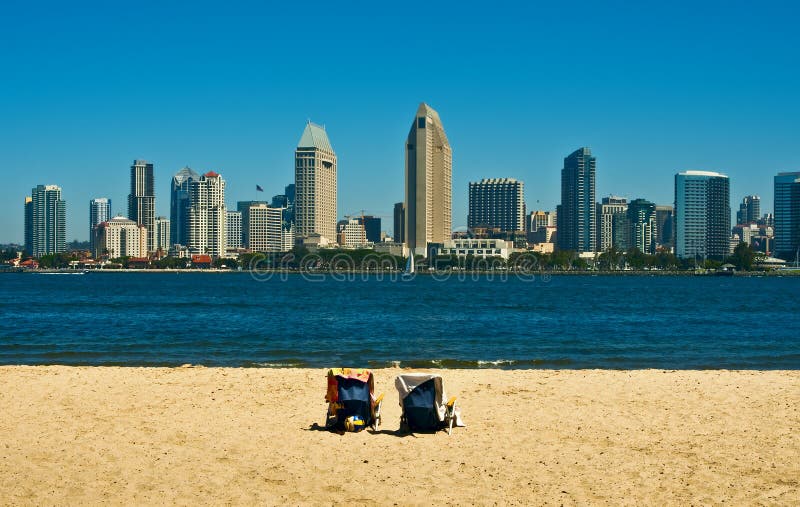 Two beach chairs, covered with towels, on the sand positioned to view the downtown San Diego skyline from Coronado, across the bay, on a beautiful sunny blue sky and water day in southern California. Two beach chairs, covered with towels, on the sand positioned to view the downtown San Diego skyline from Coronado, across the bay, on a beautiful sunny blue sky and water day in southern California.