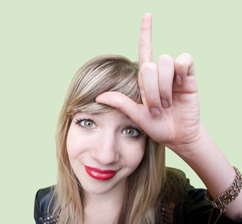 Cute young Caucasian woman makes loser sign on her forehead over green background. Cute young Caucasian woman makes loser sign on her forehead over green background