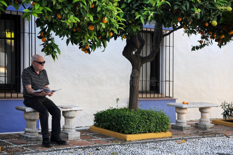 Senior man resting under orange trees and reads a magazine. He enjoys his holiday after lockdown. Senior man resting under orange trees and reads a magazine. He enjoys his holiday after lockdown