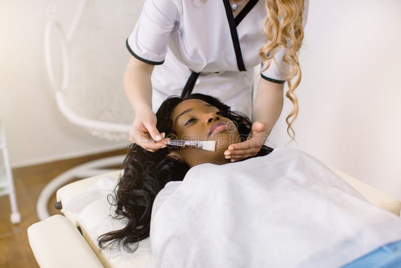 Подрезанное образ женского врача cosmetologist с помощью кисть с лечением маски кожи на лице женщины пациентов. Счастливый африкан стоковое фото rf