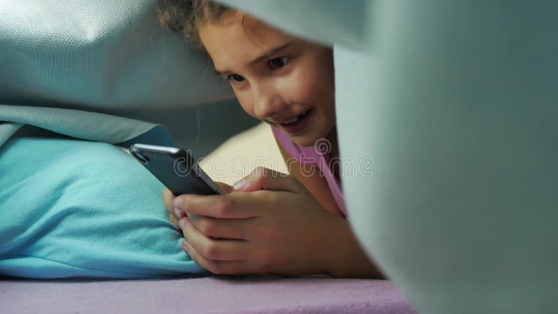 Подросток девушки и собака ночи под одеялом играя в Smartphone маленькая девочка перед кроватью лежит в кровати занимаясь серфинг видеоматериал - Видео насчитывающей темнота, ноча: 104327406->