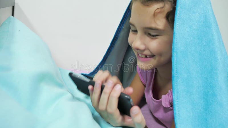 Nastolatki pod kocem bawią się w smartfonie Mała dziewczynka przed przykrytym łóżkiem leży w łóżku surfując na i stock wideo - Odtwarzanie liczenia wideo, tablet: 102383923  