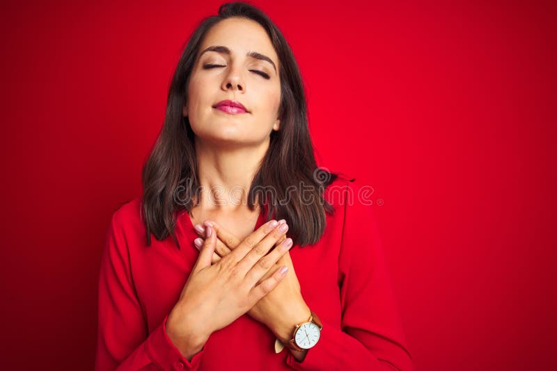 Young beautiful woman wearing shirt standing over red isolated background smiling with hands on chest with closed eyes and grateful gesture on face. Health concept. Young beautiful woman wearing shirt standing over red isolated background smiling with hands on chest with closed eyes and grateful gesture on face. Health concept