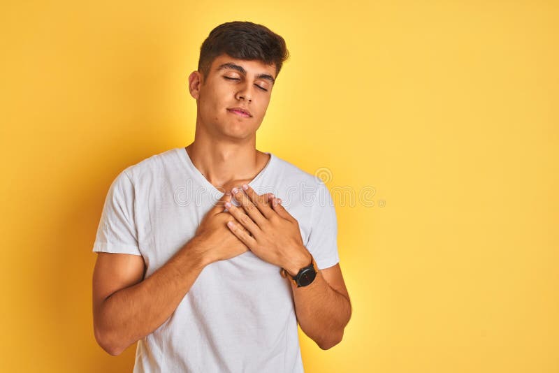 Young indian man wearing white t-shirt standing over isolated yellow background smiling with hands on chest with closed eyes and grateful gesture on face. Health concept. Young indian man wearing white t-shirt standing over isolated yellow background smiling with hands on chest with closed eyes and grateful gesture on face. Health concept