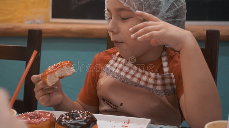Пончик красивого мальчика портрета и жевательные сладкий с укусами поливой карамельного в пекарне Закрыть поднимающий вверх мальч акции видеоматериалы