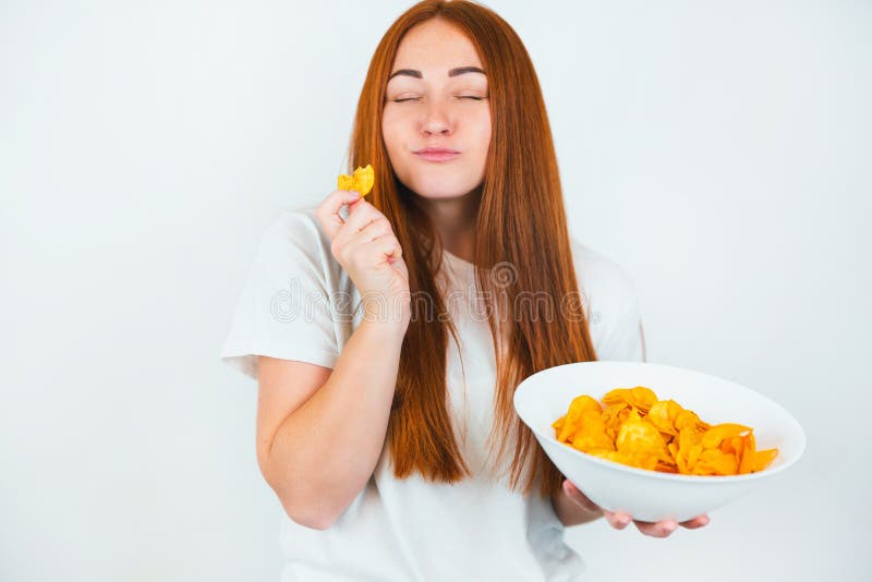 Портрет redheaded молодой женщины с укусами хрустящие картофельные чипсы с удовольствием выглядит счастливой на изолированном бело стоковые фотографии rf