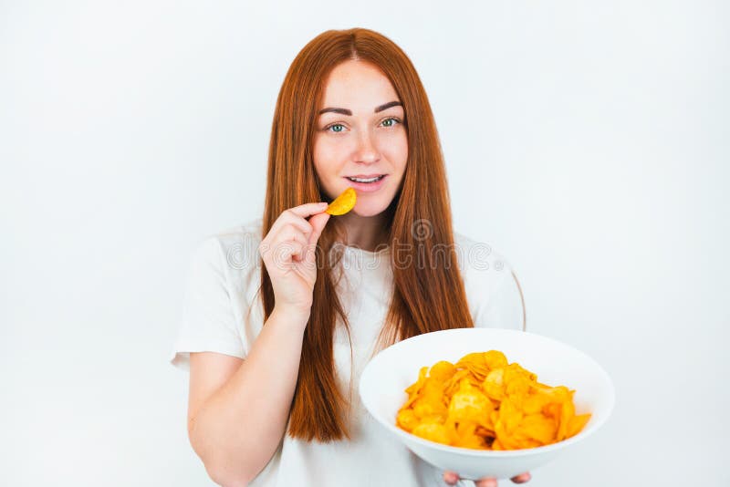 Портрет redheaded молодой женщины с укусами хрустящие картофельные чипсы выглядит счастливой на изолированной белой концепции высо стоковое изображение rf