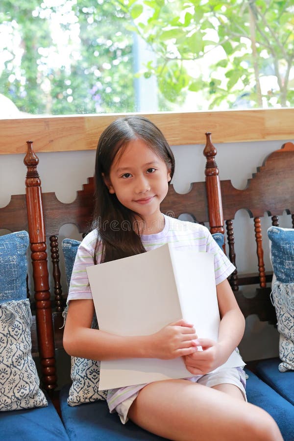 портрет азиатской девочки сидит на деревянном стуле, обняв книгу и смотря на камеру стоковое фото