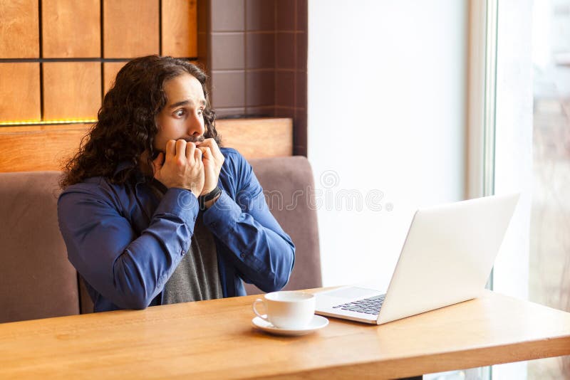 Portrait of scared handsome young adult man freelancer in casual style sitting in cafe and watching horrors movie in laptop, bite nails, bussinessman in office. Indoor, lifestyle concept. Portrait of scared handsome young adult man freelancer in casual style sitting in cafe and watching horrors movie in laptop, bite nails, bussinessman in office. Indoor, lifestyle concept