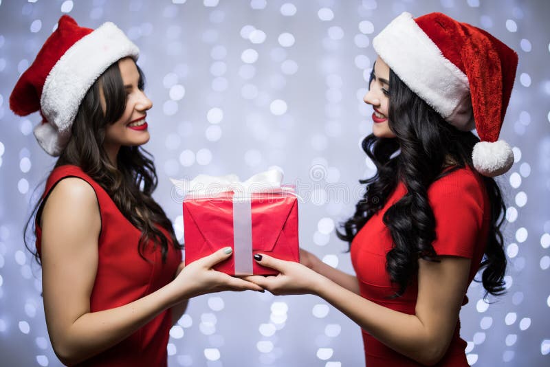 Портрет 2 женщин в шляпе santa и красное платье с подарком в руках смотрят один другого на предпосылке света bokeh Зимний отдых C стоковое изображение rf Портрет 2 женщин в шляпе santa и красное платье с подарком в руках смотрят один другого на предпосылке света bokeh Зимний отдых C стоковое изображение rf