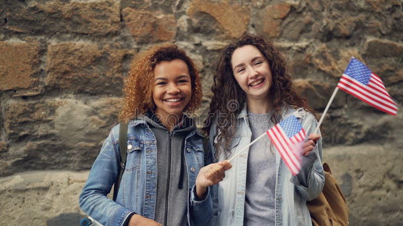 Портрет замедленного движения 2 молодых женщин друзей в вскользь одеждах развевая американские флаги и смеясь над смотрящ камеру