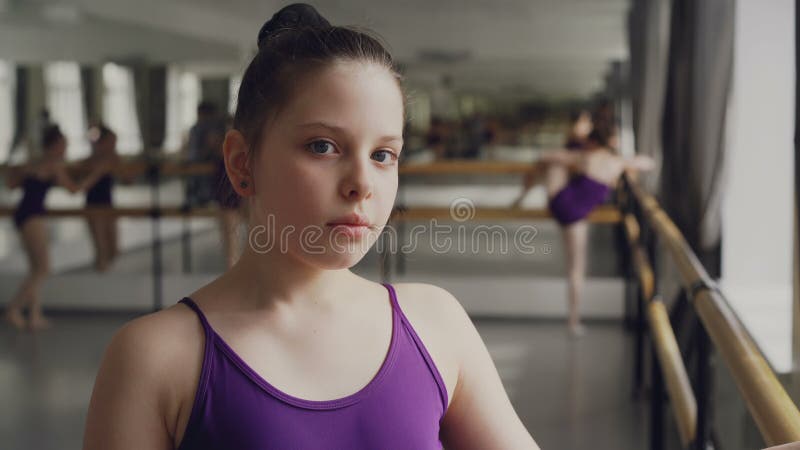 Портрет конца-вверх темн-с волосами маленькой девочки в bodysuit стоя в классе балета и смотря камеру Другие студенты