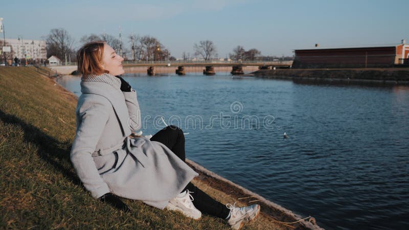 Портрет красивой женщины в сером пальто.. Молодая женщина сидит на траве возле воды и вид на панораму города весны