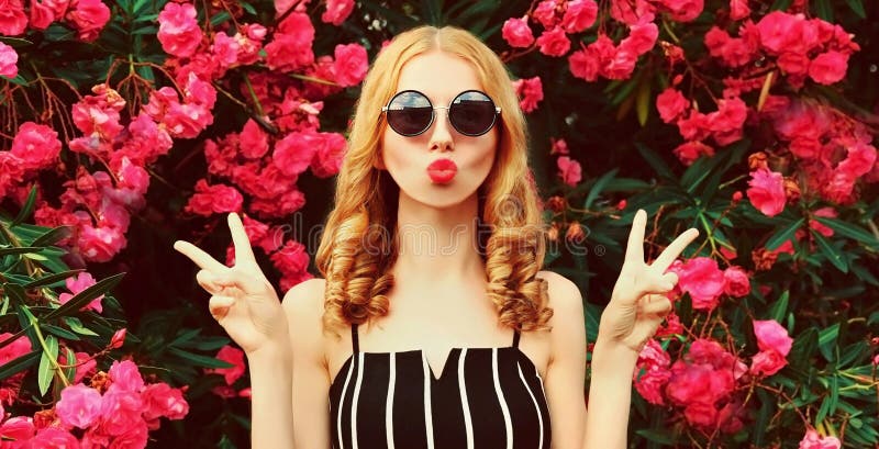 Portrait of beautiful woman blowing her red lips sending sweet air kiss on pink roses flowers background. Portrait of beautiful woman blowing her red lips sending sweet air kiss on pink roses flowers background.