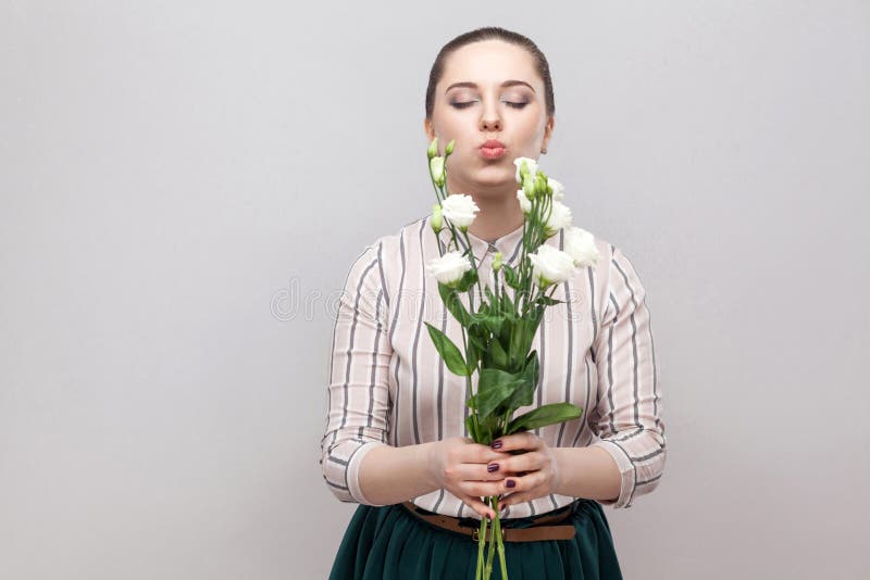 Portrait of attractive romantic lovely young woman in striped shirt and green skirt holding bouquet of white flowers and sending air kiss. Indoor, studio shot, on gray background, copy space. Portrait of attractive romantic lovely young woman in striped shirt and green skirt holding bouquet of white flowers and sending air kiss. Indoor, studio shot, on gray background, copy space