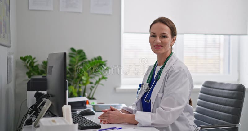 Портрет улыбающейся женщины-врача в белом халате, которая сложила руки и сидит за белым столом в современном офисе больницы сток-видео
