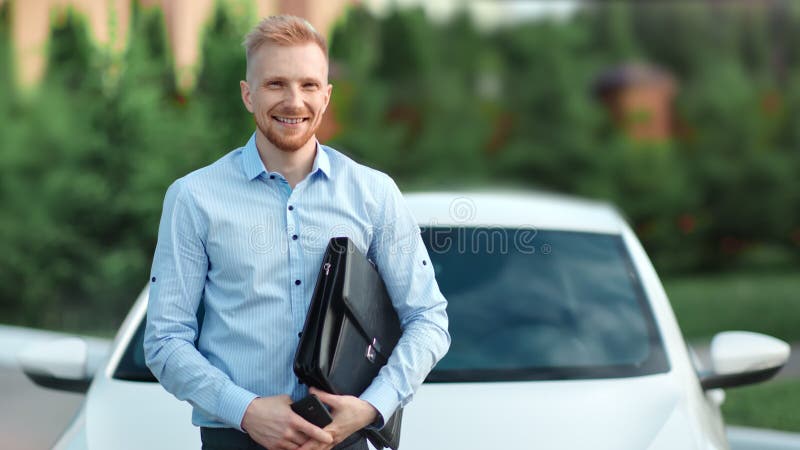 Портрет успешного усмехаясь молодого портфеля удерживания бизнесмена представляя на белой предпосылке автомобиля