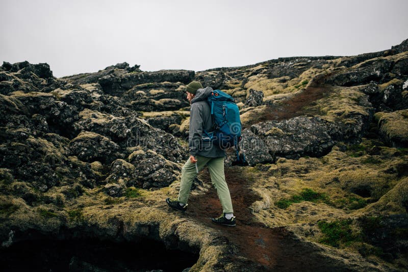 Young man in hiking trekking gear, waterproof jacket, green knit beanie and hike backpack walk through moss covered rough iceland terrain. Explore travel real wilderness lifestyle. Young man in hiking trekking gear, waterproof jacket, green knit beanie and hike backpack walk through moss covered rough iceland terrain. Explore travel real wilderness lifestyle