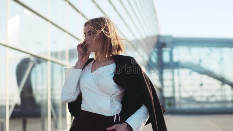 Привлекательная белокурая женщина идя около делового центра и говоря на телефоне Стильный взгляд, элегантная белая блузка