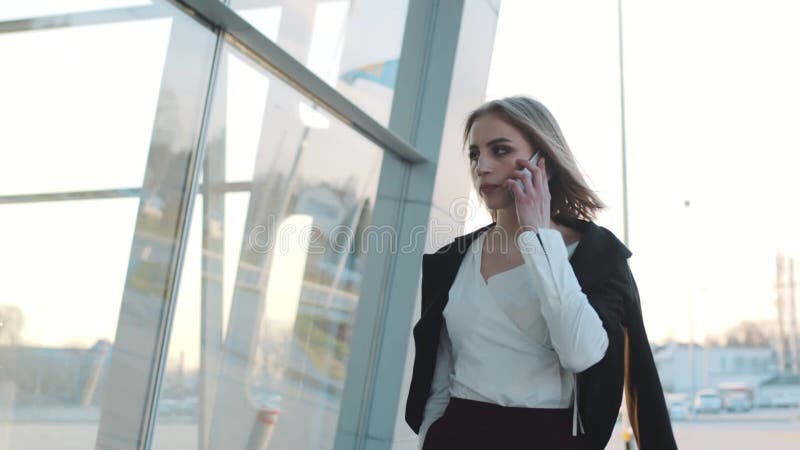 Привлекательная молодая белокурая женщина вытягивая ее чемодан и говоря на телефоне Стильное обмундирование, официально носка Был